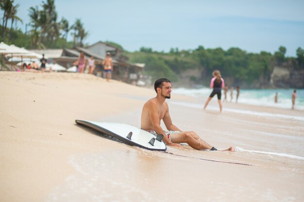 ragazzo sta riposando sulla spiaggia tropicale di sabbia dopo aver guidato il surf. Stile di vita sano e attivo nella vocazione estiva