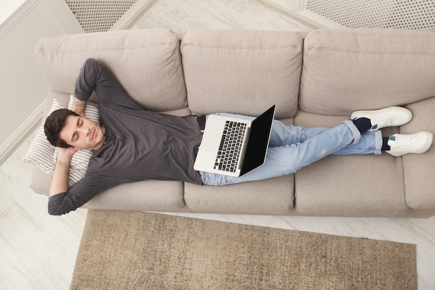 Ragazzo stanco che si riposa, libero professionista oberato di lavoro con laptop che sogna sul divano di casa, vista dall'alto, copia spazio