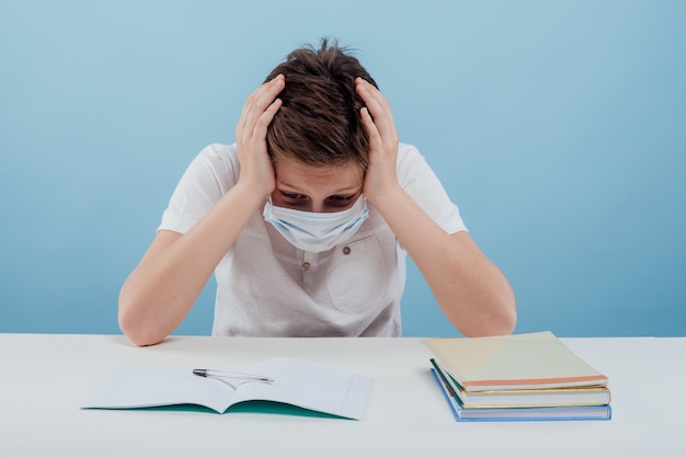 Ragazzo stanco con maschera medica le mani sulla testa seduto al tavolo isolato su sfondo blu