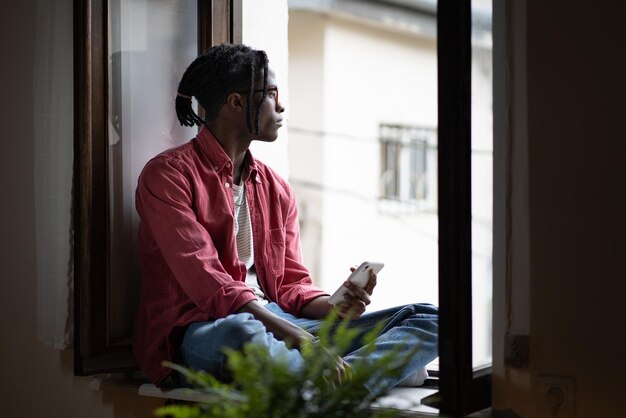 Ragazzo studente africano seduto sul davanzale della finestra che tiene gli amici scomparsi dello smartphone mentre studiava all'estero