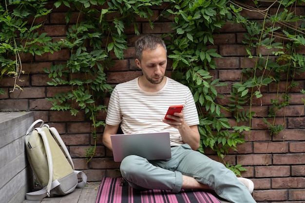 Ragazzo studente europeo seduto con il laptop all'aperto utilizzando lo smartphone distratto dal telefono cellulare