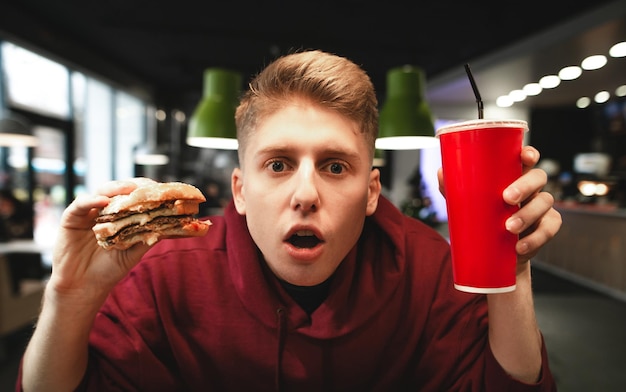 Ragazzo stupito con fast food che mangia nelle mani dello sfondo del ristorante