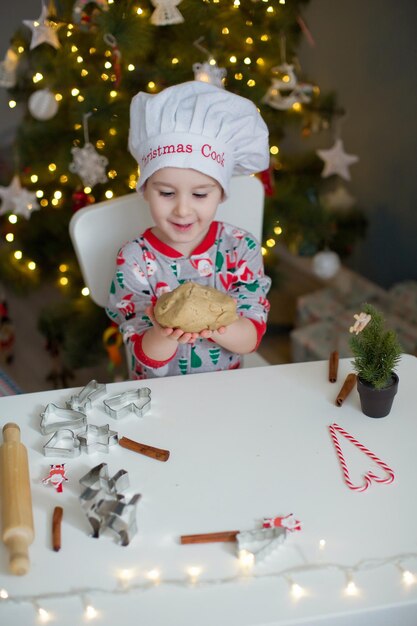 Ragazzo sveglio del bambino che fa i biscotti di Natale su un tavolo bianco vicino all'albero di Natale con le luci Concetto di cottura di Natale