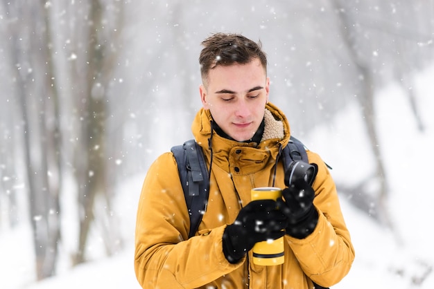 Ragazzo turistico congelato biondo che indossa una giacca arancione riscaldata da un tè caldo da una tazza termica nella fredda foresta innevata d'inverno
