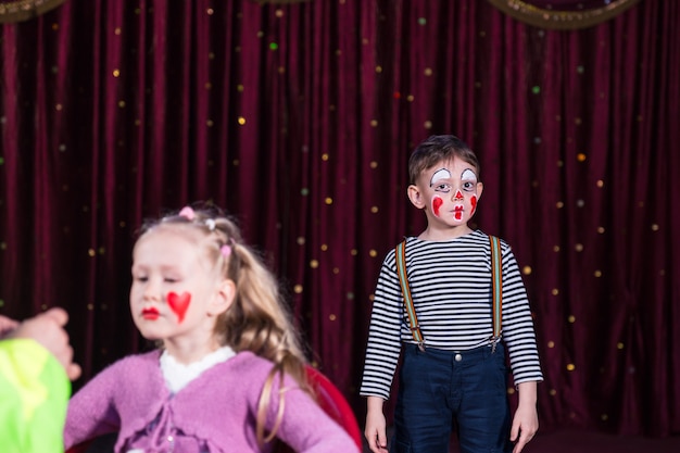 Ragazzo vestito da pagliaccio che indossa trucco e camicia a righe in piedi sul palco con sipario rosso, ragazza con trucco applicato sul viso in primo piano