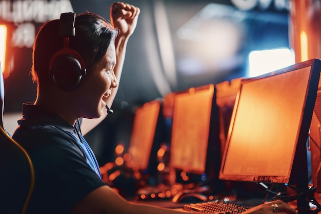 Raggiungere il successo. Vista laterale del giovane ragazzo asiatico eccitato, giocatore professionista di cybersport maschio che indossa le cuffie alzando la mano, vincendo nel torneo di eSport