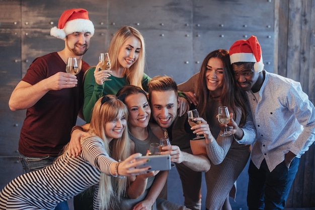 Raggruppa i bei giovani che fanno selfie nella festa di Capodanno, le ragazze e i ragazzi dei migliori amici che si divertono insieme, posando le persone di stile di vita emotivo. Cappelli di Babbo Natale e bicchieri di champagne nelle loro mani