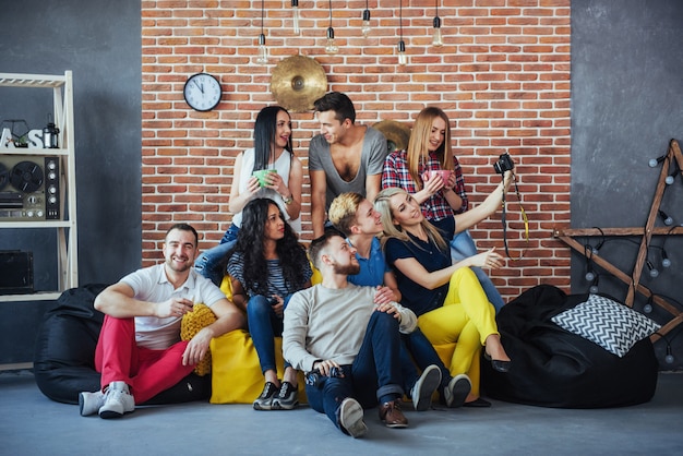 Raggruppi il ritratto dei ragazzi e delle ragazze multietnici con i vestiti alla moda variopinti che tengono l'amico che posa su un muro di mattoni, gente urbana di stile che si diverte, s circa lo stile di vita di insieme della gioventù