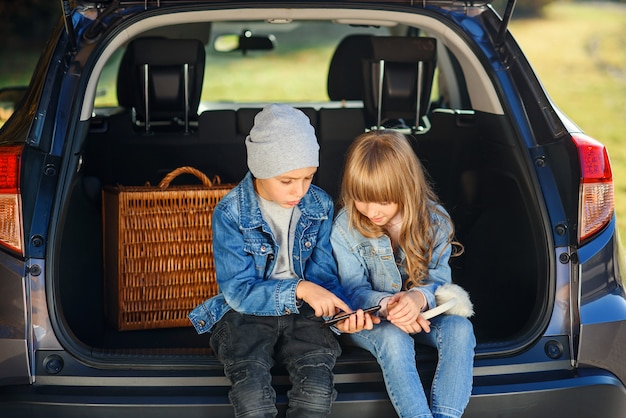 Rallentatore di ragazza bionda di 12 anni e bel ragazzo di 10 anni in jeans vestiti che seduto nel bagagliaio e guardando sul suo smartphone