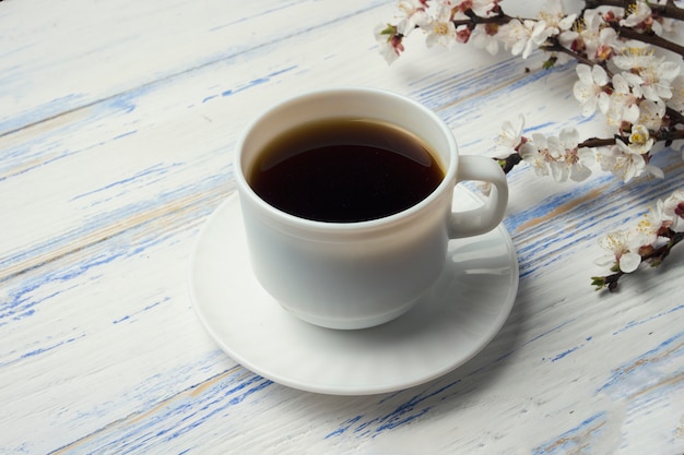 Rametto di ciliegie con fiori e tazza bianca con caffè nero su un fondo di legno bianco.