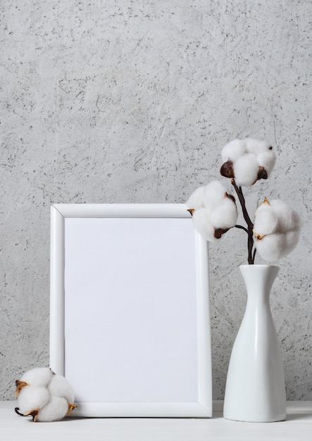 Ramo di soffici fiori di cotone in vaso bianco e cornice di legno bianca con foglio di carta vuoto su superficie di legno sopra parete chiara. Composizione floreale interna.