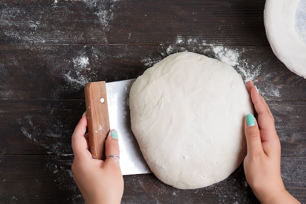 Raschietto fresco della taglierina della pasta e della taglierina della tenuta della mano della donna per la preparazione del pane casalingo su un fondo scuro di legno.