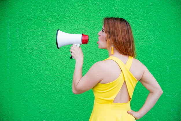 Razhevolosaya bella donna in abito giallo in piedi indietro e gridando in un megafono La ragazza fa un annuncio sull'altoparlante sullo sfondo di un muro verde Vista posteriore