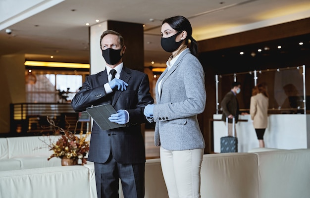 Receptionist maschi e femmine che seguono le precauzioni di sicurezza e indossano maschere stando in piedi nella hall dell'hotel