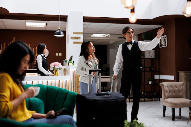 Receptionista maschio asiatico che dà indicazioni turistiche alla stanza nel corridoio di un lussuoso resort. Donna seduta nella sala salotto dell'hotel a bere caffè, in attesa di fare la prenotazione della stanza