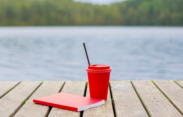 Red tazza di caffè e notebook posa sul molo in legno sullo sfondo del lago