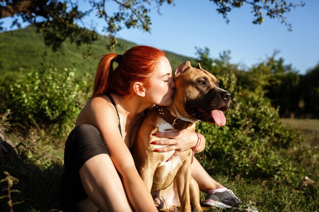 Redhair jouful giovane donna che accarezza il loro cane indossando abbigliamento sportivo godendo il loro tempo e le vacanze nel soleggiato parco