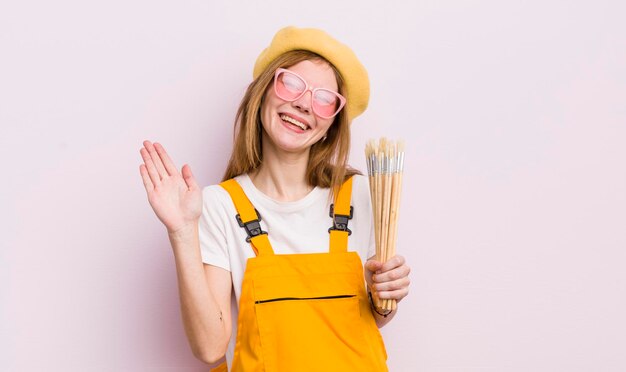 Redhead bella ragazza sorridente felicemente agitando la mano dandoti il benvenuto e salutandoti concetto di studente d'arte