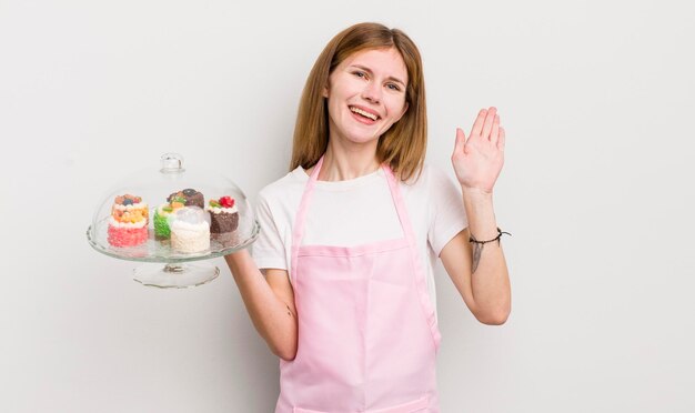 Redhead bella ragazza sorridente felicemente agitando la mano dandoti il benvenuto e salutandoti concetto di torte fatte in casa