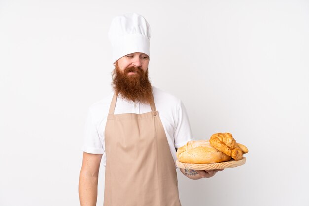 Redhead in uniforme da chef. Panettiere maschio che tiene una tabella con parecchi pani con l'espressione triste