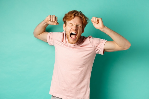 Redhead maschio fan guardando la partita di sport e tifo, gridando e facendo pompe a pugno, guardando la telecamera, guardando la concorrenza, tifando per la squadra, in piedi su sfondo turchese.