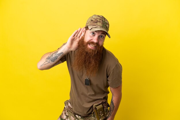 Redhead militare con dog tag isolato su sfondo giallo ascoltando qualcosa mettendo la mano sull'orecchio