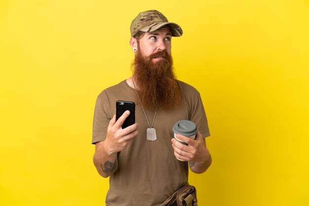 Redhead militare con dog tag isolato su sfondo giallo tenendo il caffè da portare via e un cellulare mentre pensa a qualcosa