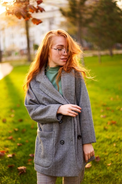 Redhead sorridente bella donna che cammina nel parco in una soleggiata giornata autunnale Elegante posa femminile in un abito elegante e autentico si diverte a ridere sul prato verde con foglie di fogliame vivide La ragazza gode della stagione autunnale