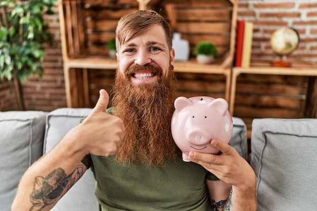 Redhead uomo che tiene salvadanaio sorridendo felice e positivo, pollice in su facendo eccellente e segno di approvazione