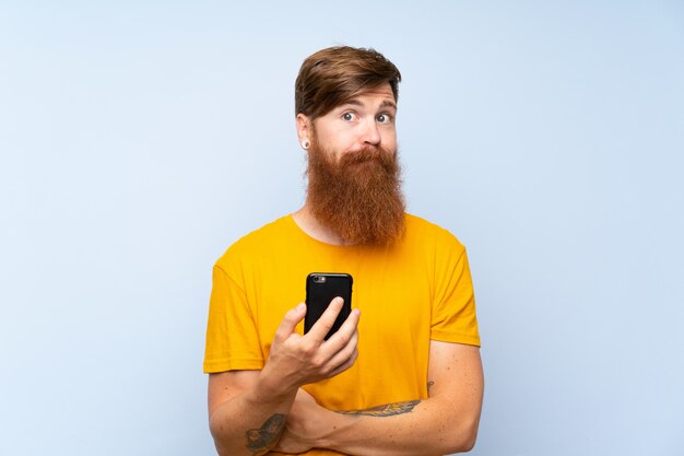 Redhead uomo con la barba lunga con un cellulare sul muro blu facendo dubbi gesto mentre si sollevano le spalle