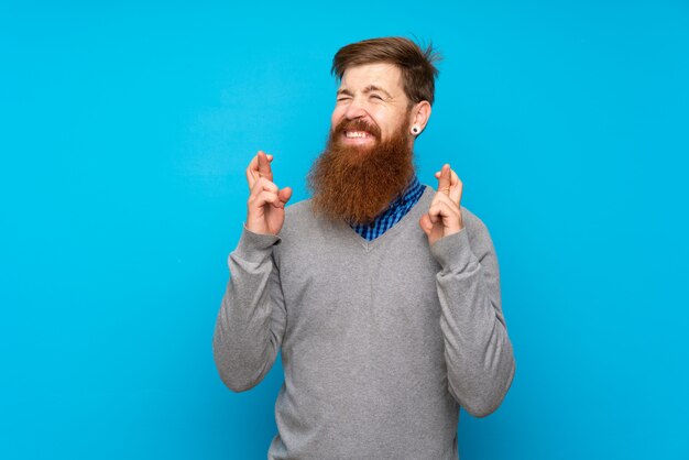 Redhead uomo con la barba lunga sopra isolato blu con le dita che attraversano e desiderano il meglio