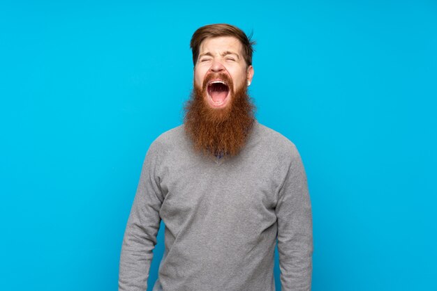 Redhead uomo con la barba lunga sopra isolato blu gridando in avanti con la bocca spalancata