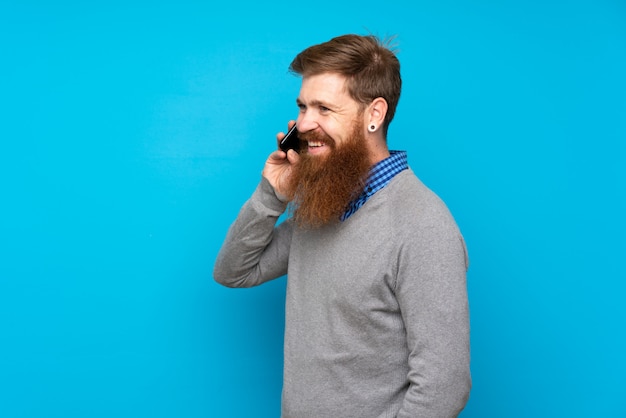 Redhead uomo con la barba lunga sul muro blu mantenendo una conversazione con il telefono cellulare