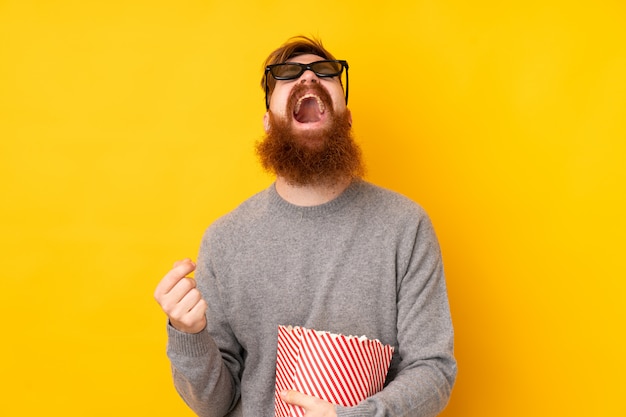 Redhead uomo con la barba lunga sul muro giallo isolato con occhiali 3d e in possesso di un grande secchio di popcorn