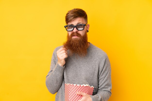 Redhead uomo con la barba lunga sul muro giallo isolato con occhiali 3d e in possesso di un grande secchio di popcorn
