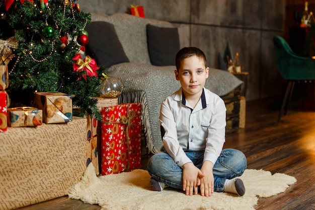 Regali di apertura del ragazzo felice durante la vigilia di Natale. Bel ragazzo seduto sul pavimento accanto albero di Natale. Kid festeggia le vacanze invernali. Il Natale è tempo di regali. Famiglia e stile di vita