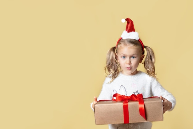 Regali di Natale di consegna. Ragazza bambino eccitato sorpreso in cappello di Babbo Natale rosso posa isolato sfondo giallo tenere scatola di cartone kraft presente. Vendita di prodotti stagionali per lo shopping online.