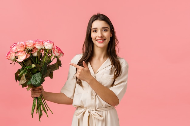 Regali, San Valentino, concetto di bellezza. La splendida fiorista femminile ha realizzato un bellissimo bouquet per il cliente, tenendo le rose e puntando il dito contro i fiori, sorridendo sciocca e felice, parete rosa