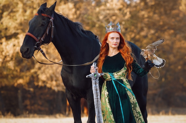 Regina con i capelli rossi in un abito verde con una corona e una spada vicino a un cavallo con un uccello