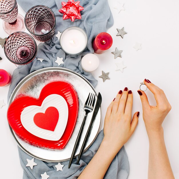 Regolazione della tavola di San Valentino. Le mani femminili tengono la fede nuziale. Piatti bianchi e rossi a forma di cuore, bicchieri per bevande, candele e posate sfondo bianco. Concetto romantico