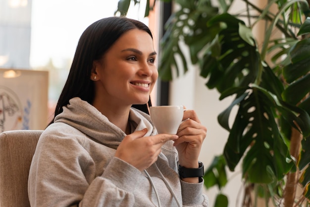 Relax mattutino. Bruna donna seduta in poltrona e godendo della sua tazza di caffè contro la finestra, foto d'archivio