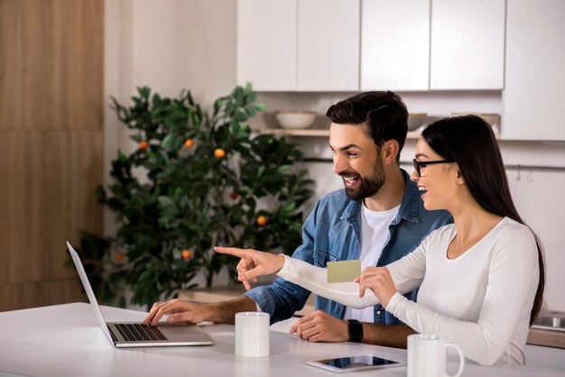 Relazioni felici Giovani coppie allegre che si siedono in cucina mentre usano il loro computer portatile