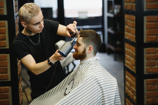 Rendere perfetto il taglio di capelli. Giovane uomo barbuto che ottiene taglio di capelli dal parrucchiere mentre era seduto in poltrona al barbiere