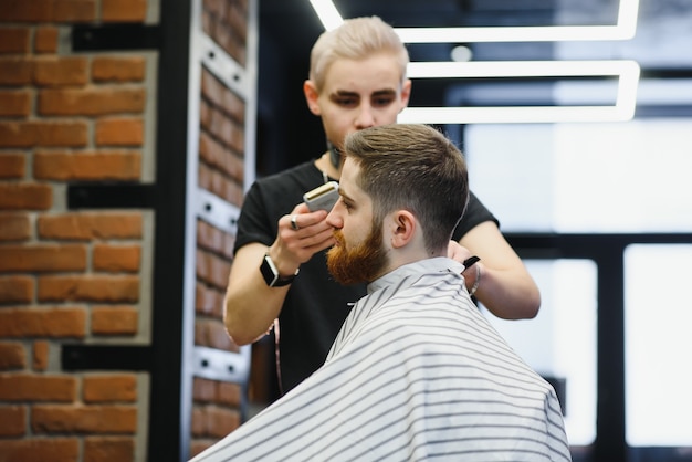 Rendere perfetto il taglio di capelli. Giovane uomo barbuto che ottiene taglio di capelli dal parrucchiere mentre era seduto in poltrona al barbiere