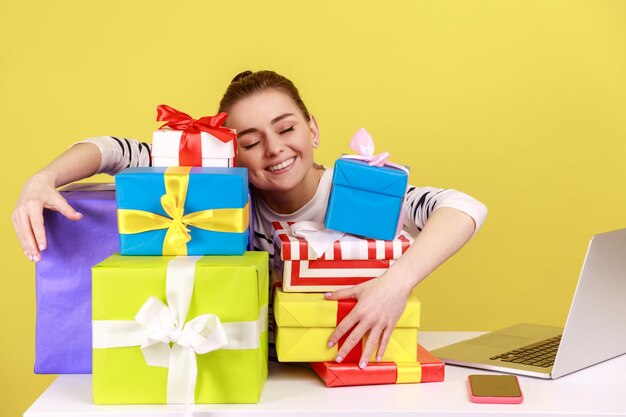 Responsabile dell'ufficio della donna soddisfatta sorridente felice che si siede sul posto di lavoro e che abbraccia molte scatole presenti