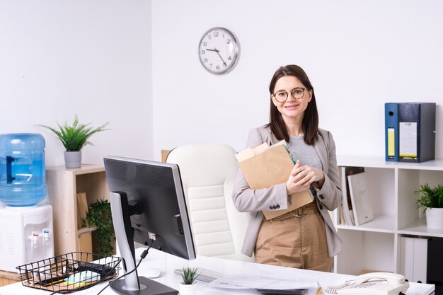 Responsabile di ufficio o contabile sorridente abbastanza giovane con i documenti che ti guarda mentre fa una pausa sul posto di lavoro con il computer