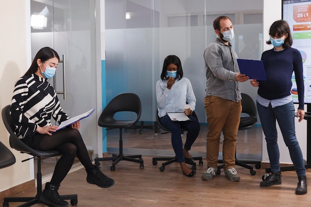 Responsabile in stato di gravidanza del dipartimento delle risorse umane che indossa una maschera protettiva che parla con l'uomo dell'offerta di lavoro mentre esamina il suo curriculum. Diverse persone in sala d'attesa che leggono i termini e le condizioni.