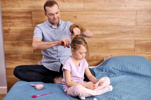 Responsabile padre papà intrecciare i capelli delle figlie, i preparativi per la scuola, bella ragazza bambino premuroso maschio seduto sul letto