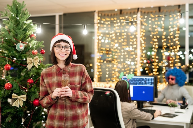 Responsabile ufficio femminile allegro con flauto di champagne in piedi da albero di Natale decorato