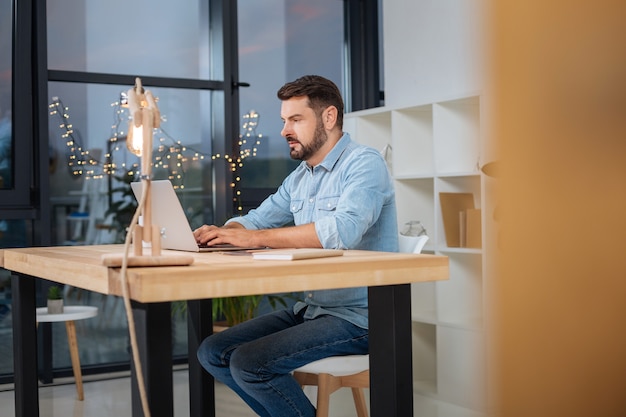 Responsabilità e duro lavoro. Uomo bello intelligente serio seduto al laptop e concentrandosi sul suo compito mentre lavora fino a tardi nel suo ufficio